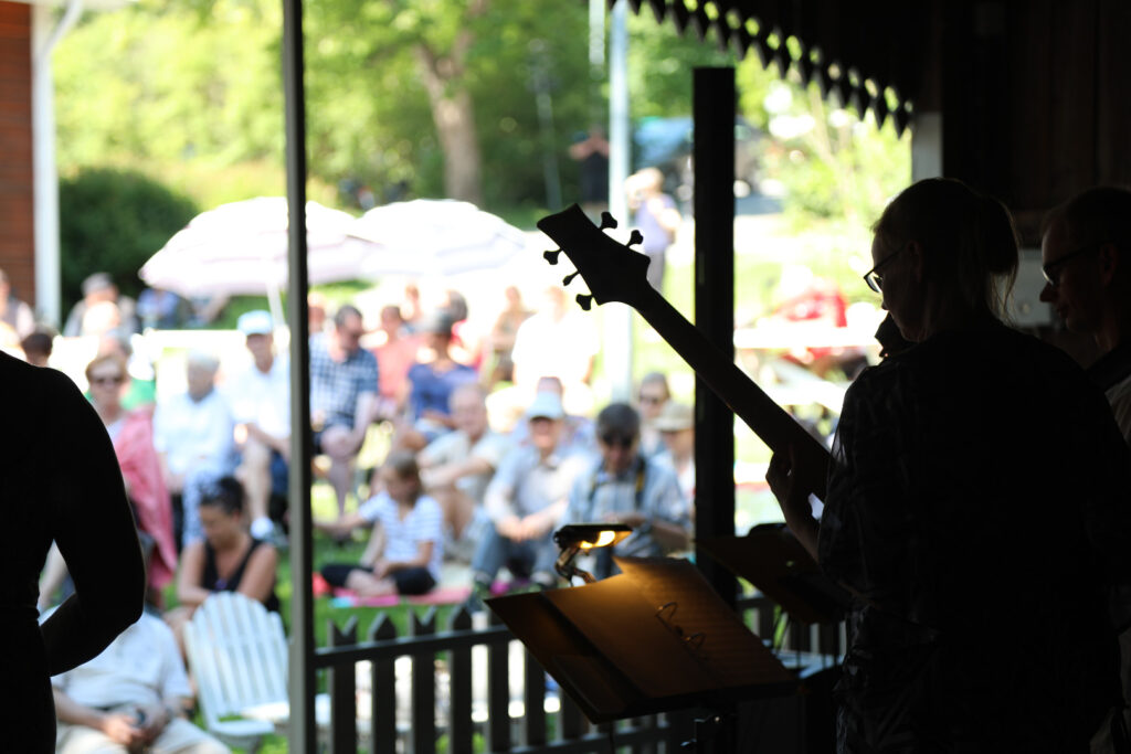 Ulkoilmakonsertissa esiintyy kitaristi, jonka siluetti piirtyy mustana. Taustalla yleisö nauttii konsertista aurinkoisessa kesäsäässä.